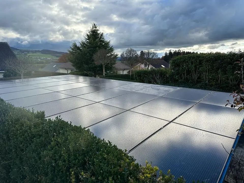 Schwarze Solarmodule auf dem grauen Ziegeldach eines Einfamilienhauses in Chénens (Umgebung: Wiesen, Bäume)